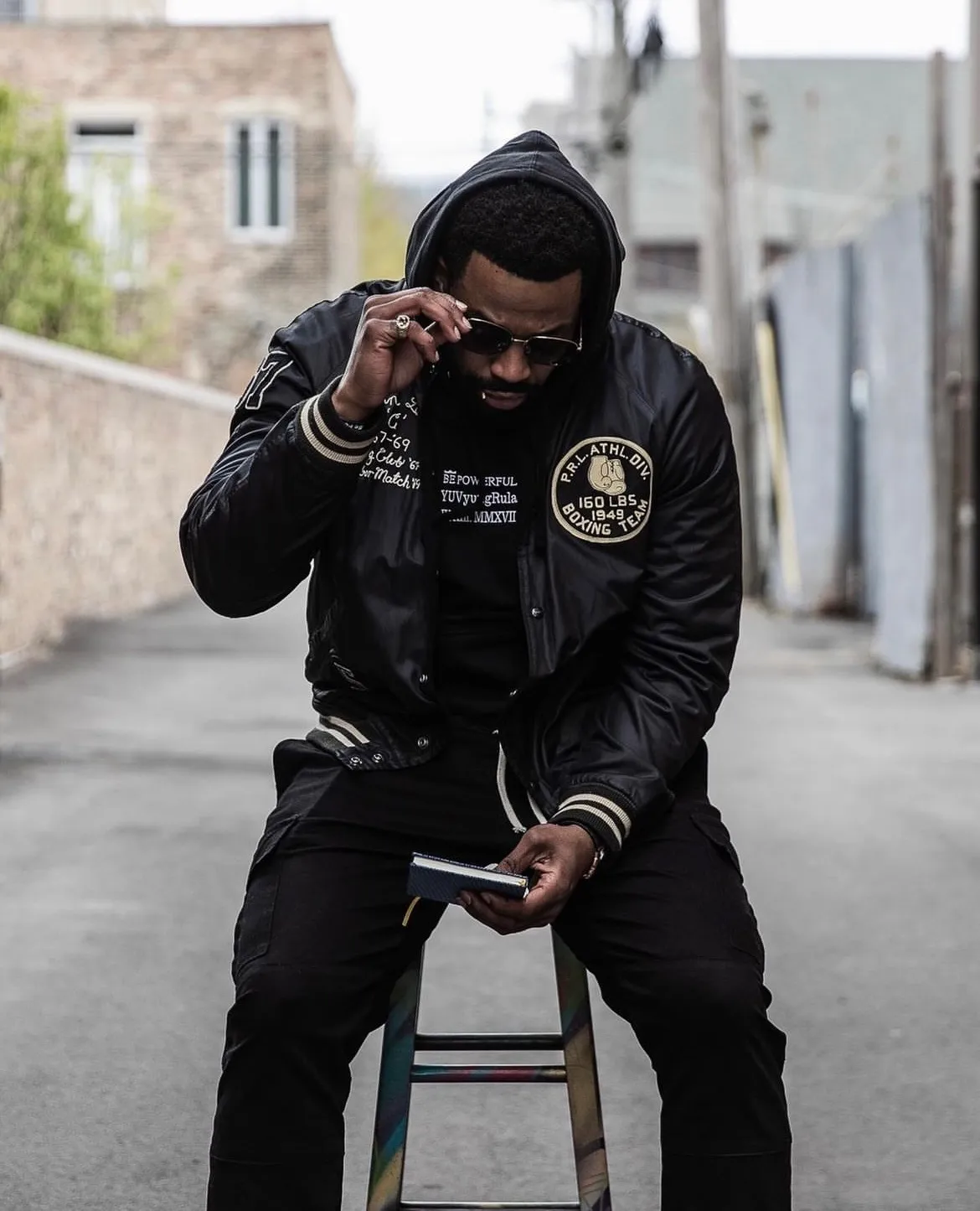 LaRoyce on a stool in an alley wearing a black varsity jacket, adjusting his sunglasses